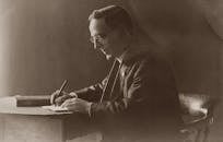 A vintage portrait of a man in a black suit writing at a desk.